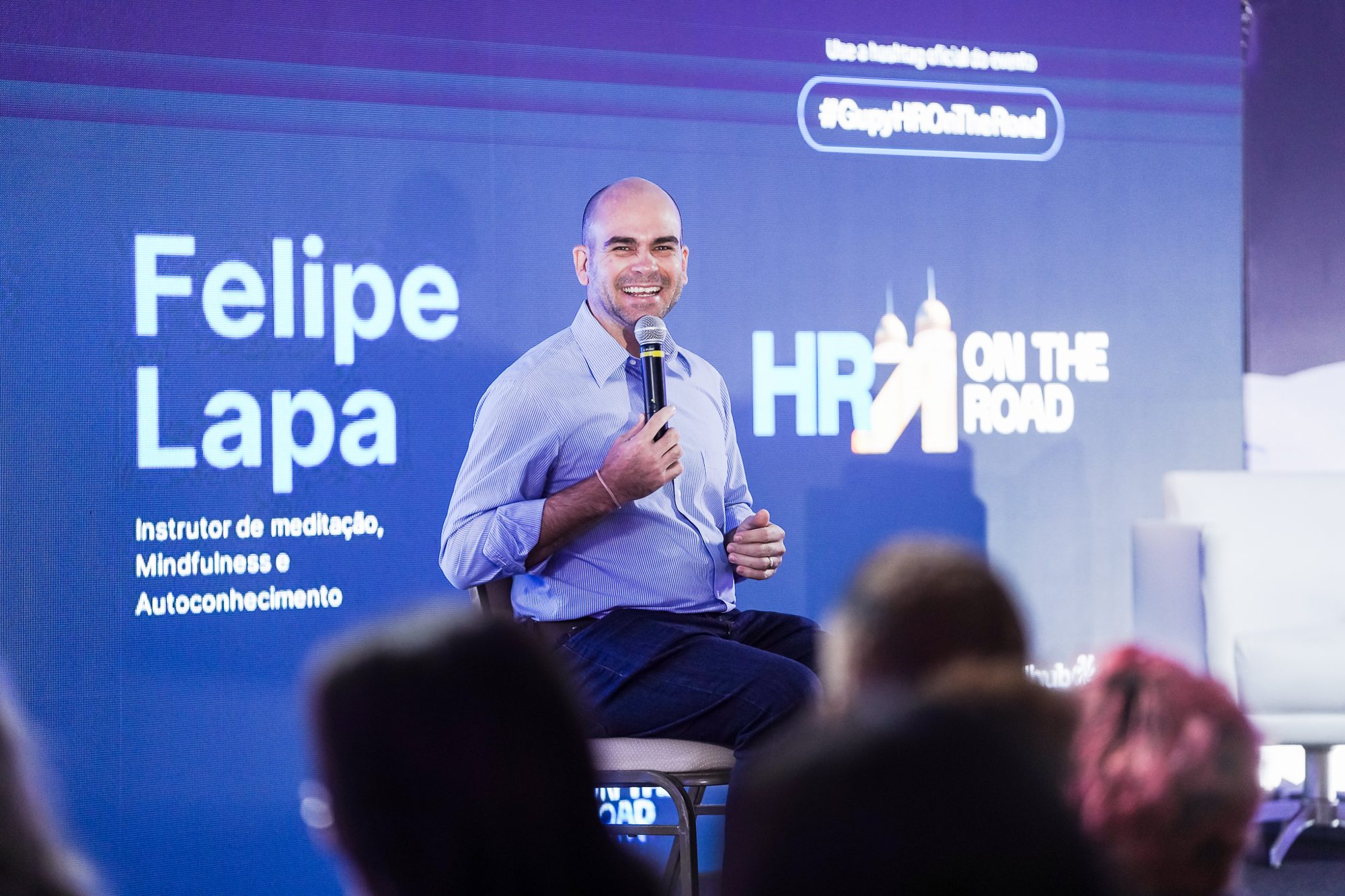 Felipe Lapa sentado no palco, ao microfone, com o nome dele no telão do HR On The Road, em Recife